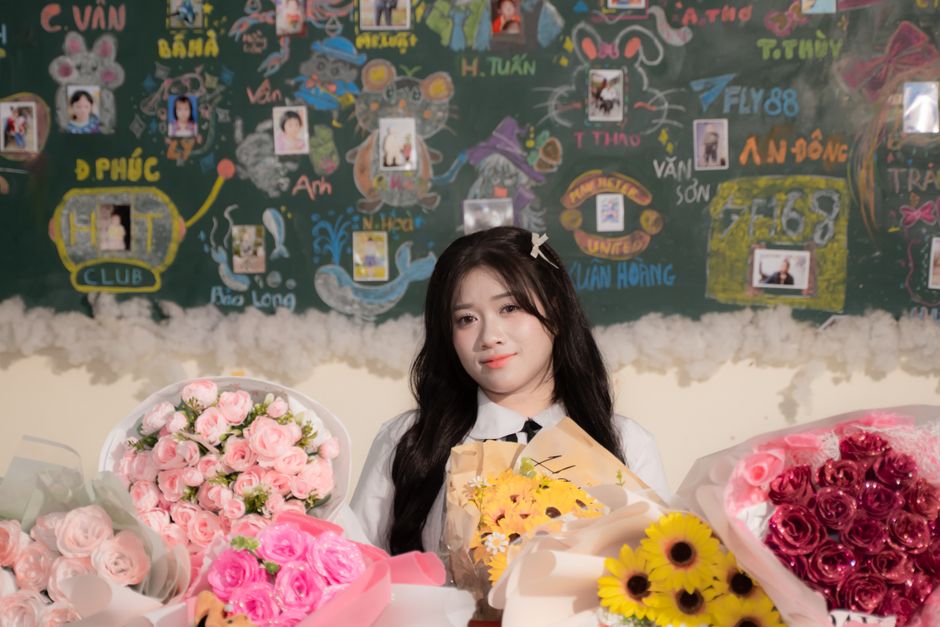 A young woman with long dark hair smiling gently while sitting behind a table of floral arrangements. She is holding a bouquet of bright yellow flowers wrapped in brown paper, with her other hand rest