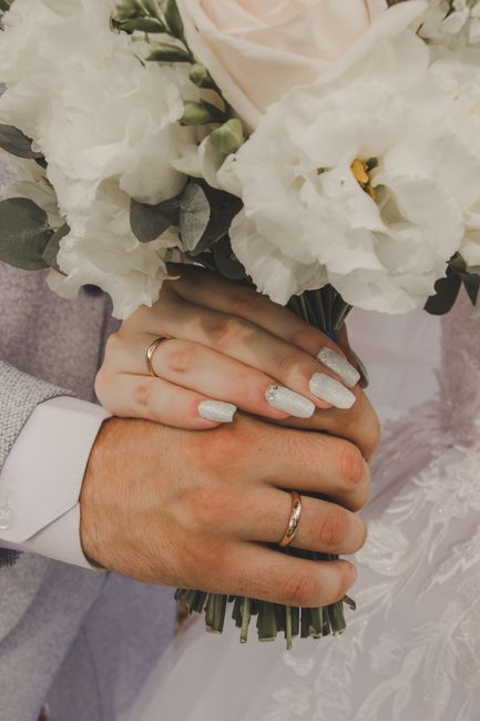 Close-up of a bride and groom's hands holding a bouquet of white flowers, including lisianthus and other delicate blooms, wrapped in dark green stems. The bride's hand displays a silver wedding band a
