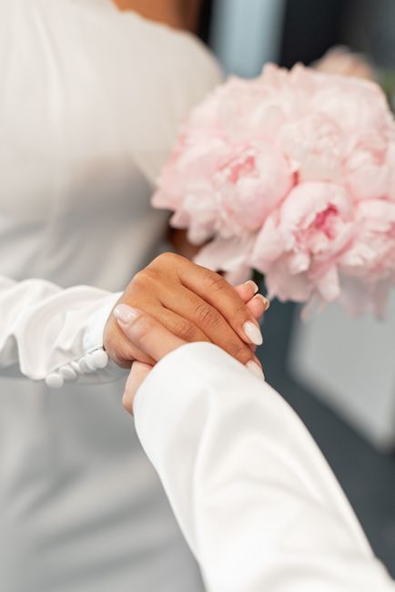 Close-up of a person in a white long-sleeved dress holding a bouquet of soft pink peonies with lush, ruffled petals. The bouquet is wrapped in clear cellophane, and the person’s hands are gently gra