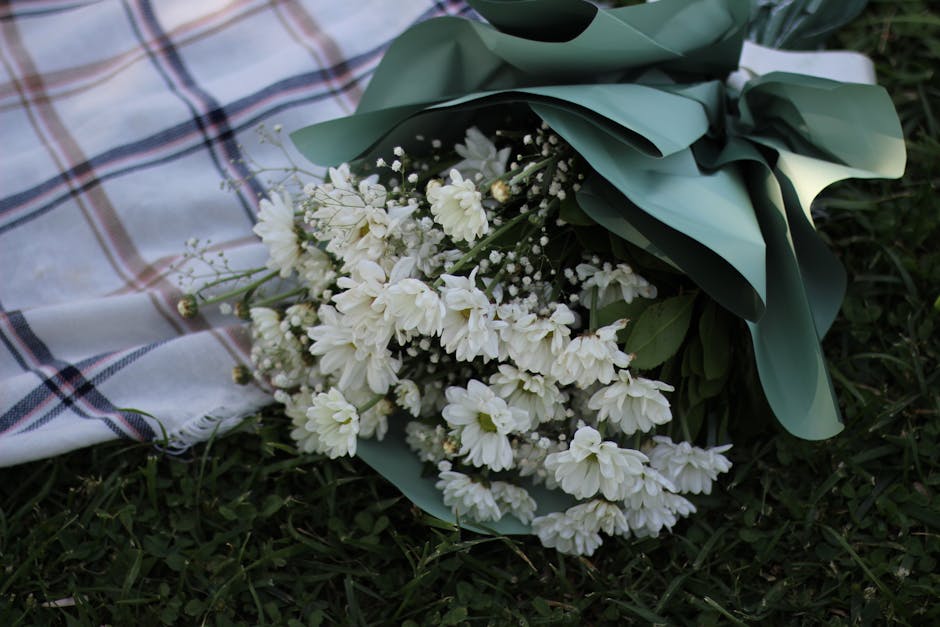 A bouquet of white flowers, including small daisies and delicate baby's breath, is arranged with lush green leaves and wrapped in pastel green paper with a white ribbon. The bouquet is placed on a gra