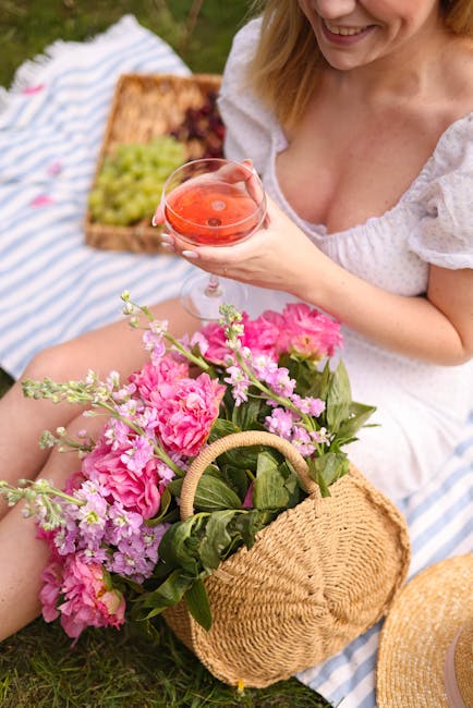 A woman sitting outdoors on a picnic blanket holds a glass of pink cocktail, smiling. In her lap, there is a wicker basket overflowing with vibrant pink peonies, purple stock, and lush green leaves, c