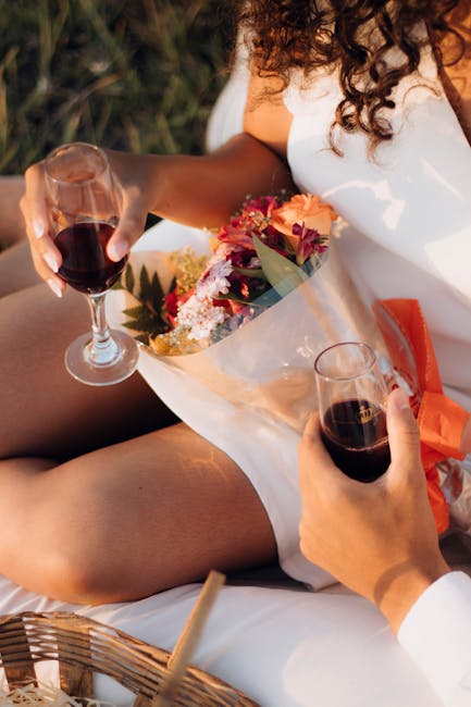 A person wearing a white dress holds a wrapped bouquet of colorful flowers, including pink, orange, and white blooms with green foliage, while sitting outdoors on a picnic blanket. One hand casually h